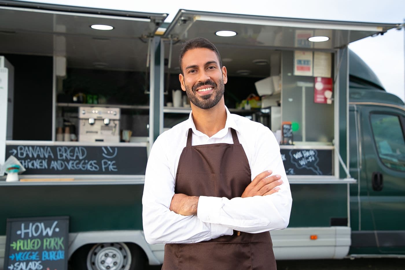 Dono de um pequeno negócio, de avental e braços cruzados, sorrindo na frente do seu estabelecimento